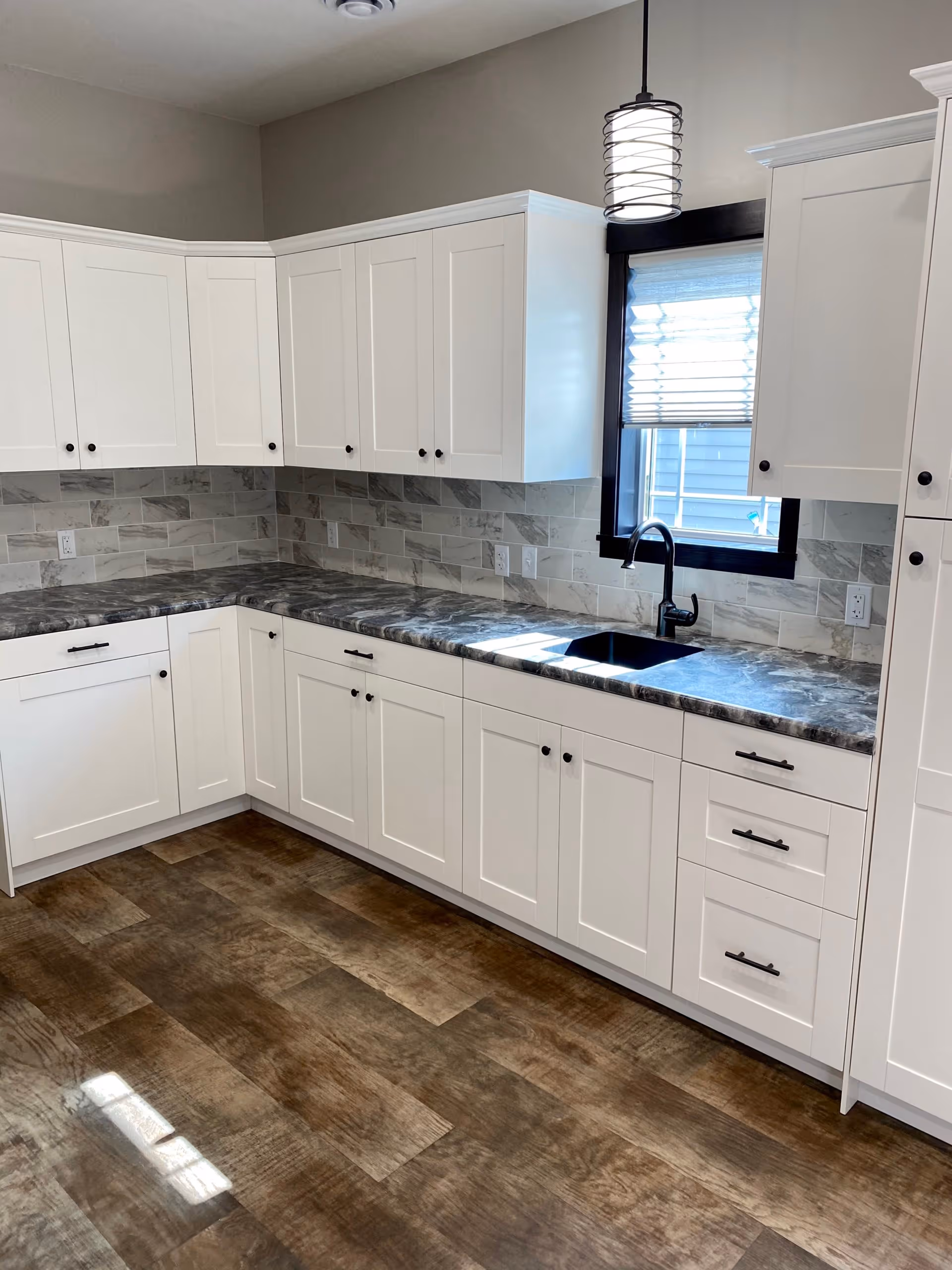 Modern kitchen with white cabinets, dark marble countertops, a black sink and faucet under a window with blinds, beige tiled backsplash, and wood-patterned flooring.