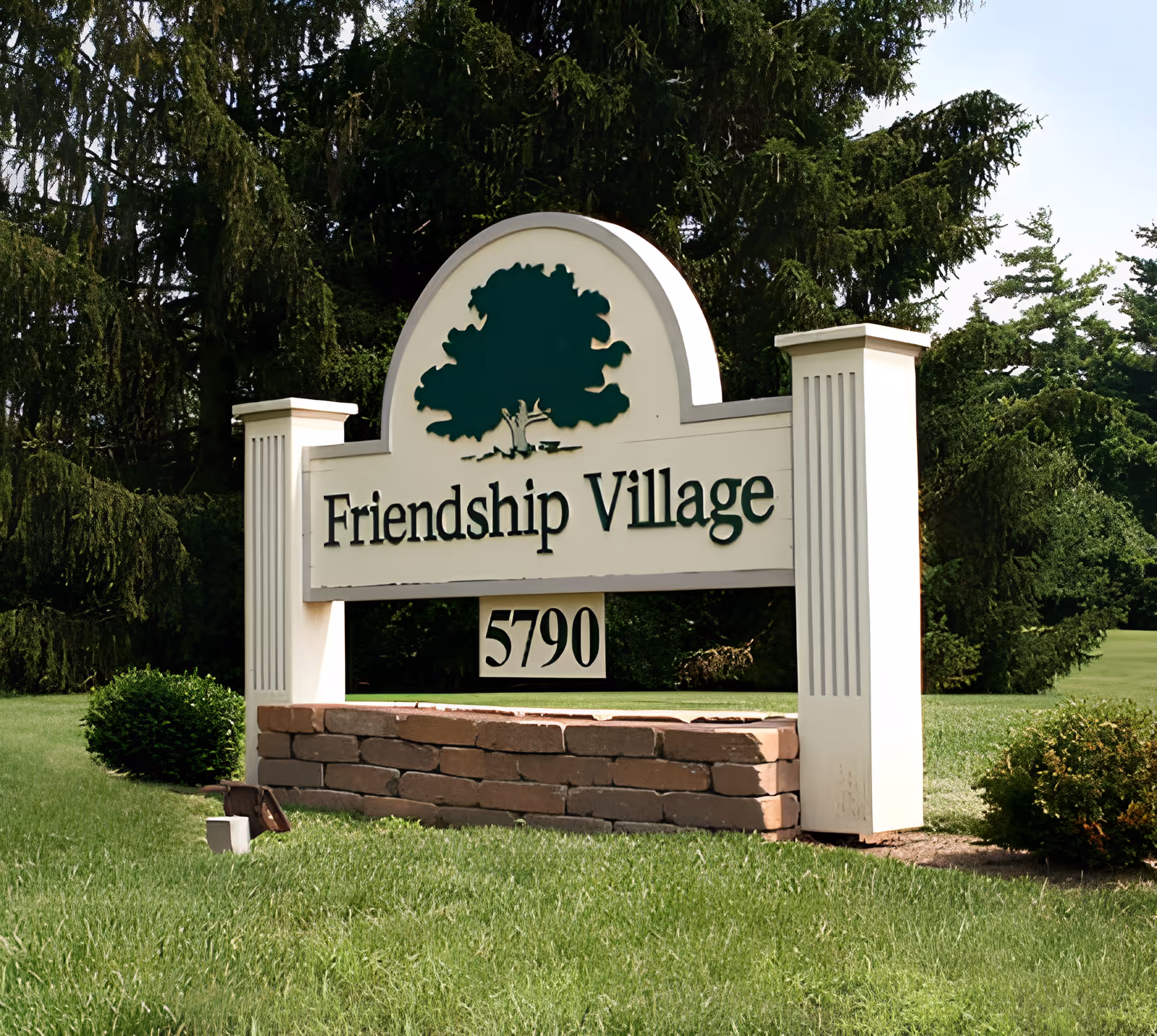 A large outdoor sign for Friendship Village with the number 5790 displayed below the name. The sign features a green tree graphic and is set on a brick base surrounded by grass and trees in the background.
