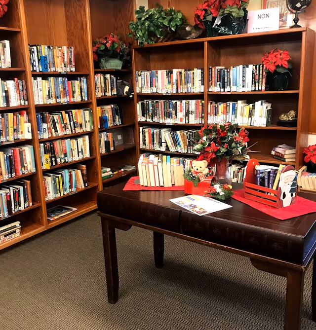 Interior view of a library room with wooden bookshelves filled with books. A wooden table in the center holds a vase with red flowers, a teddy bear, and a few other decorative items. The shelves are labeled with letters and there are green plants on top of the shelves.
