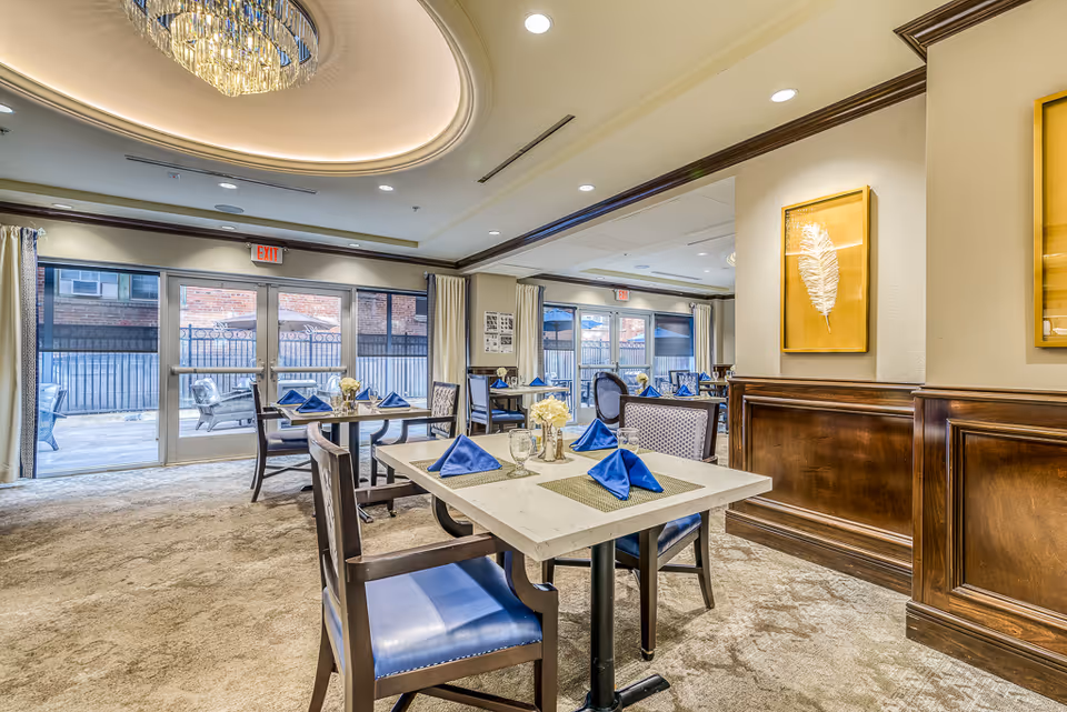 A bright and elegant dining room in a senior living facility with tables set with blue napkins, glassware, and small flower arrangements. The room features large windows and glass doors leading to an outdoor patio area with seating. The ceiling has a decorative recessed light fixture, and the walls are adorned with framed artwork. The carpeted floor and wooden paneling add warmth to the space.