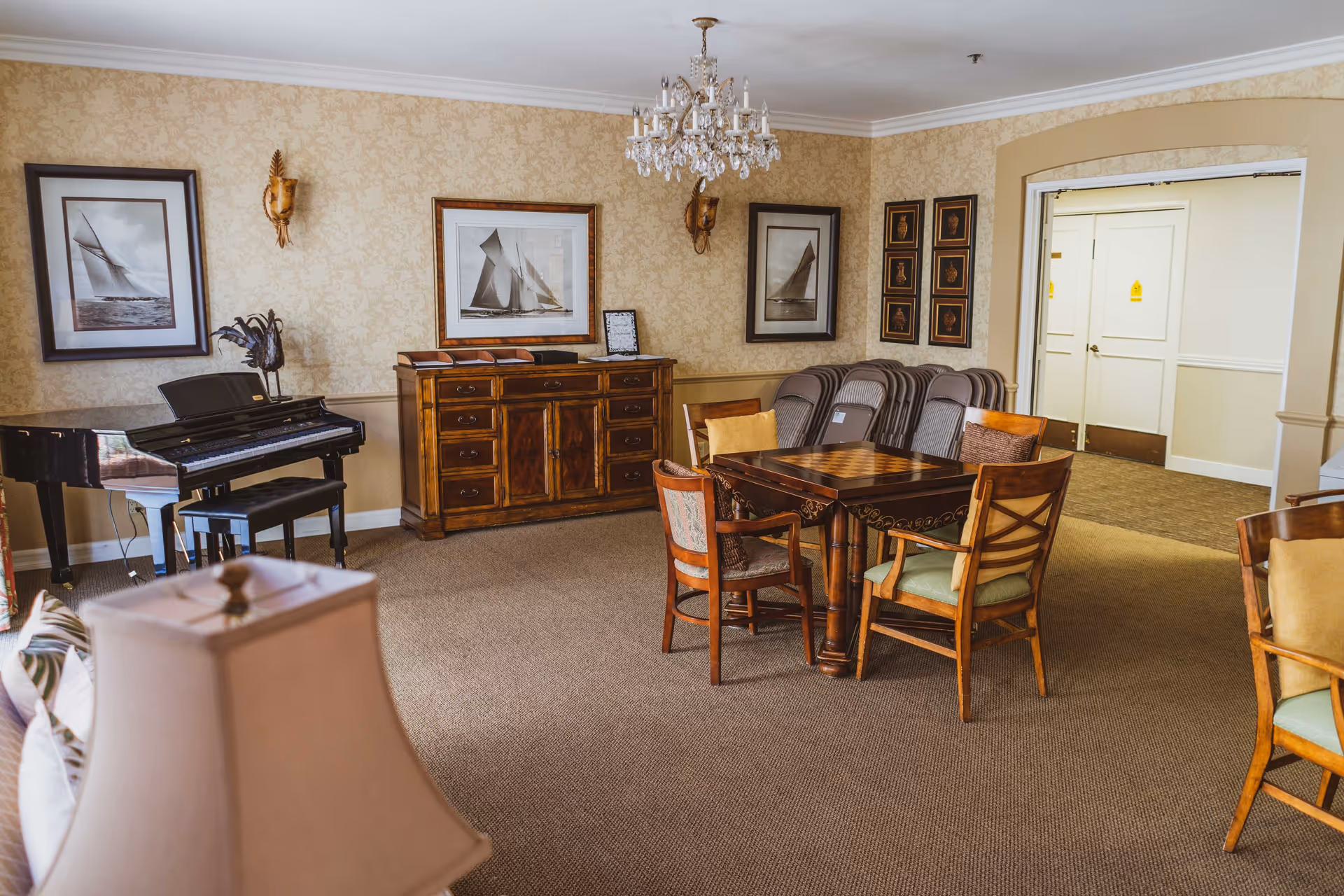 A furnished common room with a grand piano, wooden table and chairs, chandelier, and framed nautical artwork on patterned wallpaper.