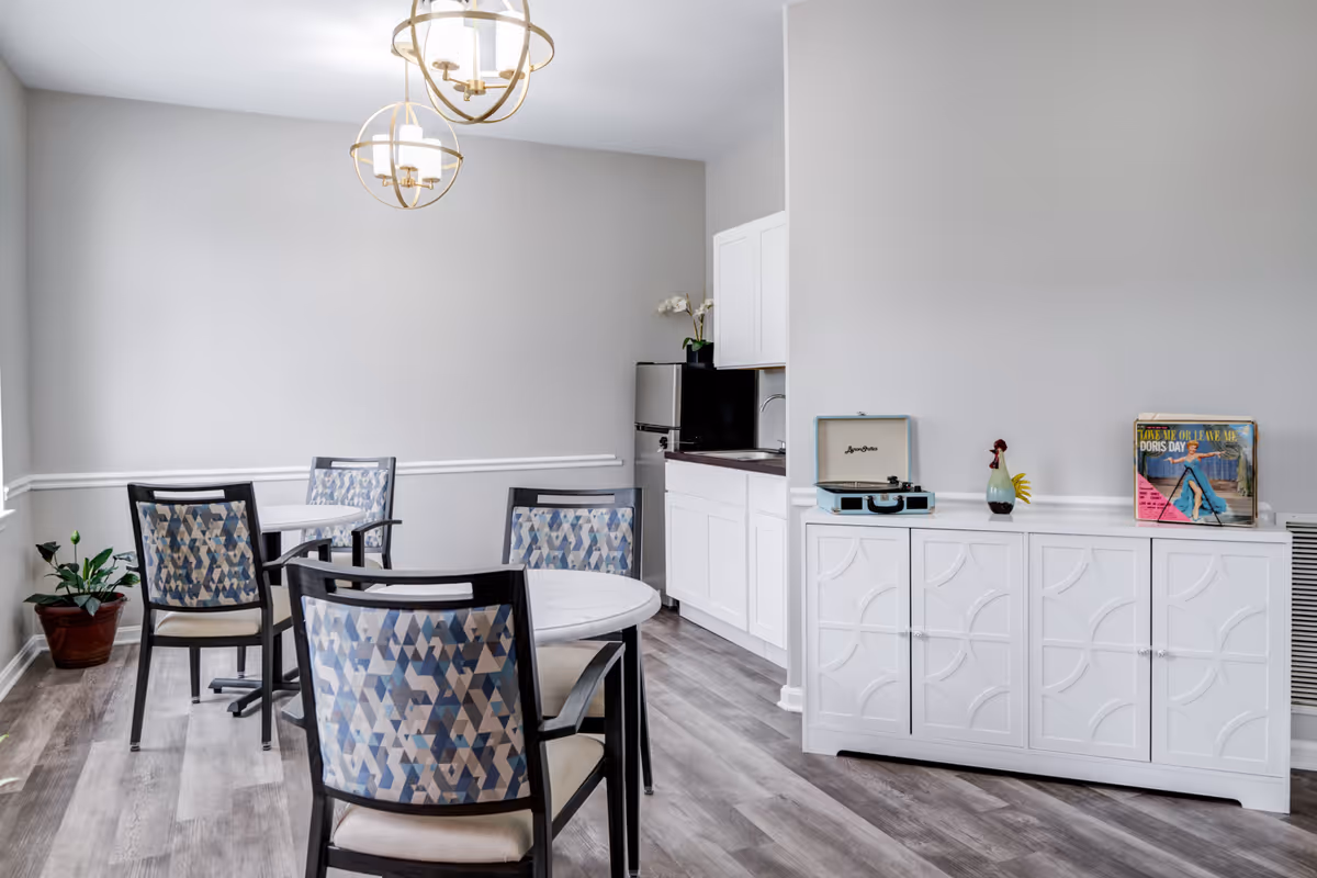A bright dining area with two round tables and patterned chairs, a small kitchenette with white cabinets and a mini fridge, and a white sideboard with decorative items including a record player and a vinyl record. The room has light gray walls, wood flooring, and two modern pendant lights hanging from the ceiling.