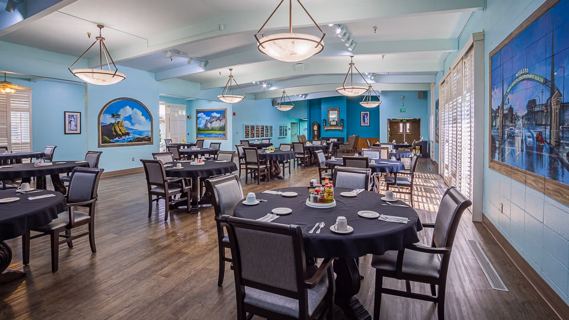 A spacious dining room with multiple round tables covered with black tablecloths, each set with white plates, cups, and silverware. The room has light blue walls adorned with large paintings and windows with white shutters allowing natural light to enter. Several hanging light fixtures illuminate the room, and wooden flooring adds warmth to the space.