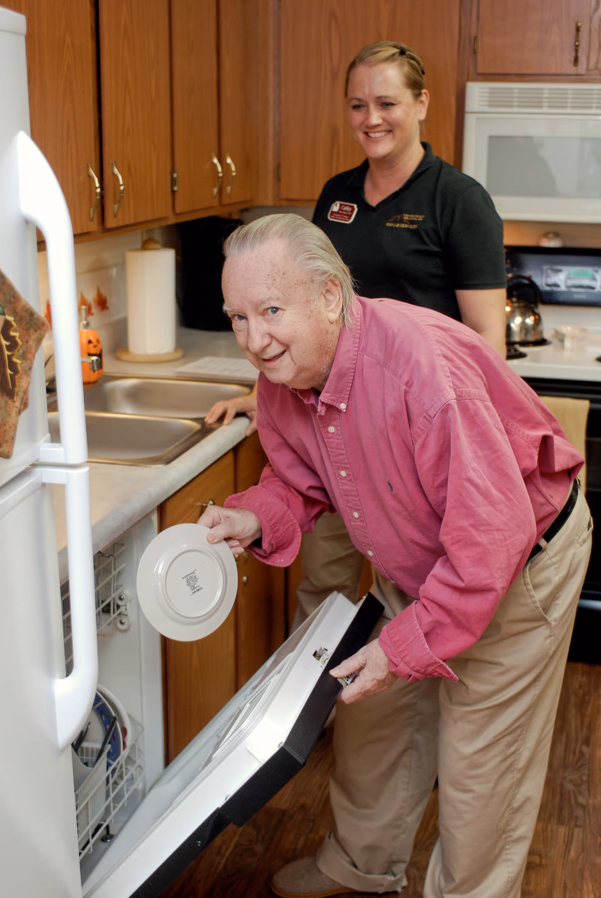 An elderly man loads a dishwasher in a kitchen while a smiling staff member stands behind him.