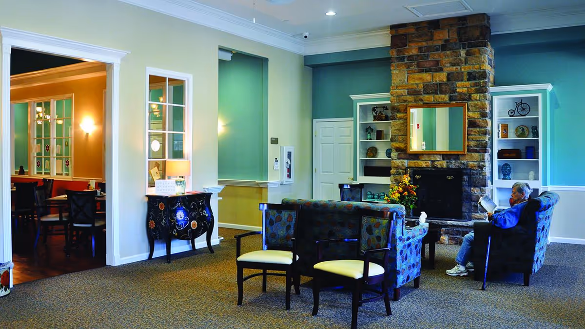 Elderly man reading in a sitting area with upholstered chairs, a stone fireplace, and built-in shelves in a senior living common room.