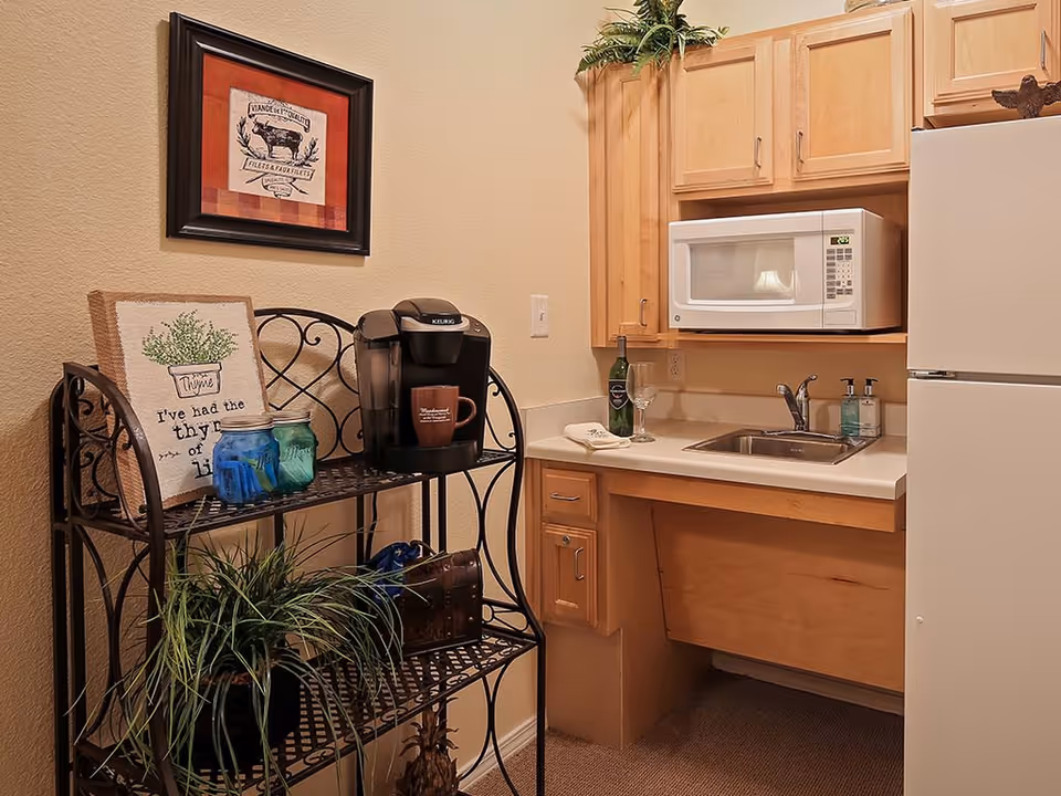 A small kitchen area with light wood cabinets, a white microwave, a white refrigerator, and a sink with two soap dispensers. To the left, there is a black metal shelf holding a Keurig coffee maker, a brown mug, two glass jars, a framed decorative sign, and a potted plant. A framed picture hangs on the wall above the shelf.