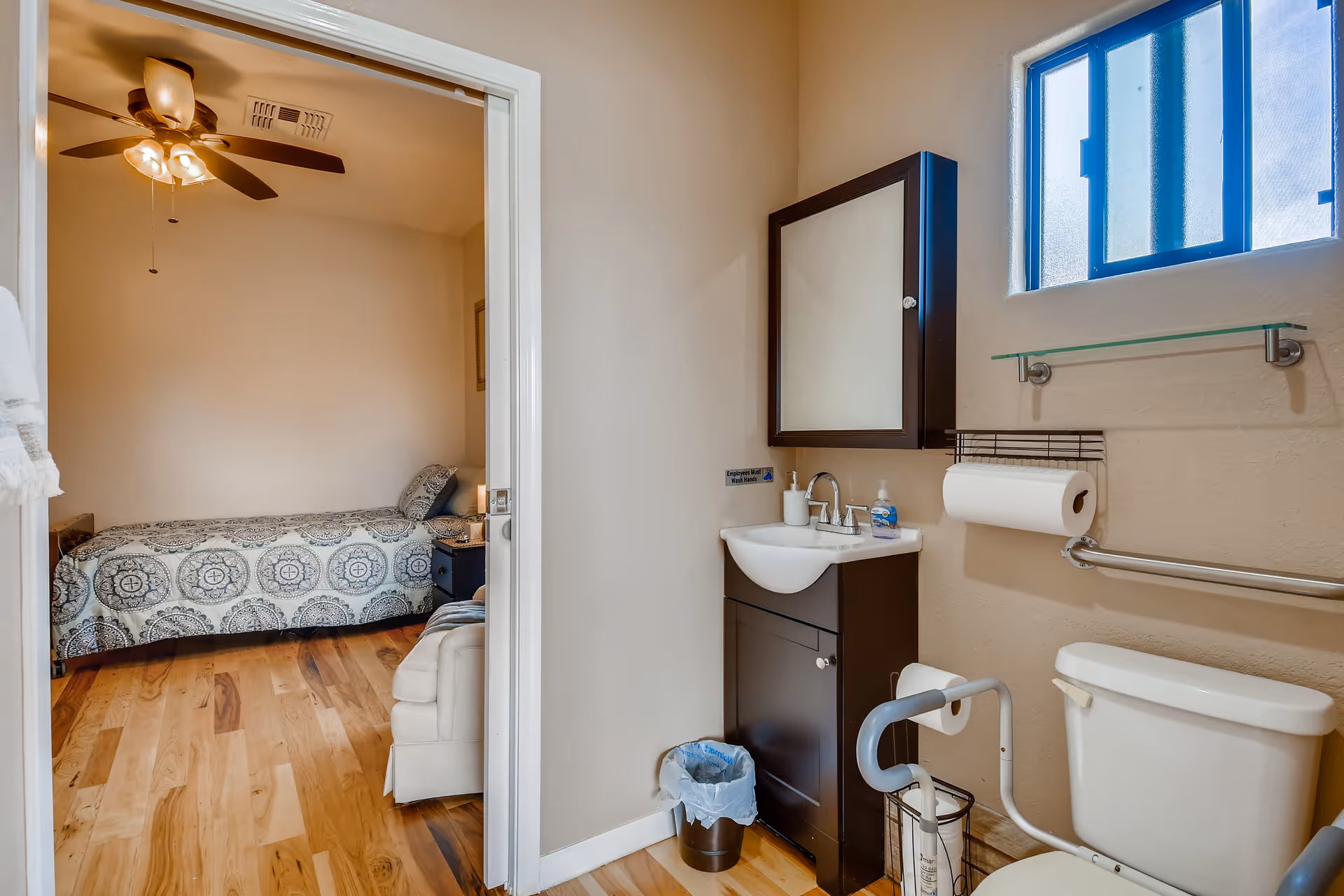 View of a small bathroom with a toilet, sink with a mirror cabinet above it, a paper towel holder, and a small window. The bathroom has beige walls and wooden flooring. Through an open door, a bedroom with a single bed, nightstand, and ceiling fan is visible.