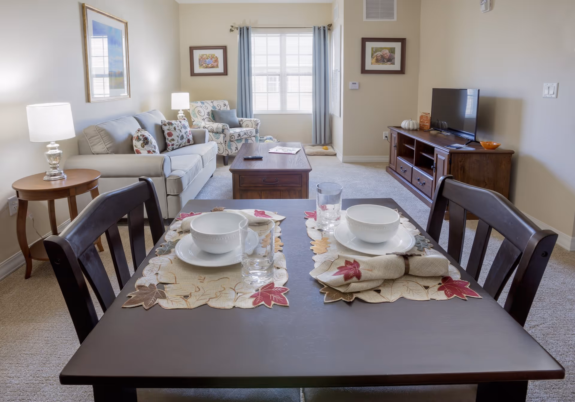 A cozy living and dining area in an assisted living facility. The foreground shows a dark wooden dining table set for two with white bowls, plates, glasses, and autumn-themed placemats and napkins. In the background, there is a beige sofa with floral pillows, a patterned armchair, a wooden coffee table, and a wooden TV stand with a flat-screen TV. The room is softly lit with a table lamp and natural light coming through a window with blue curtains. Family photos hang on the walls.
