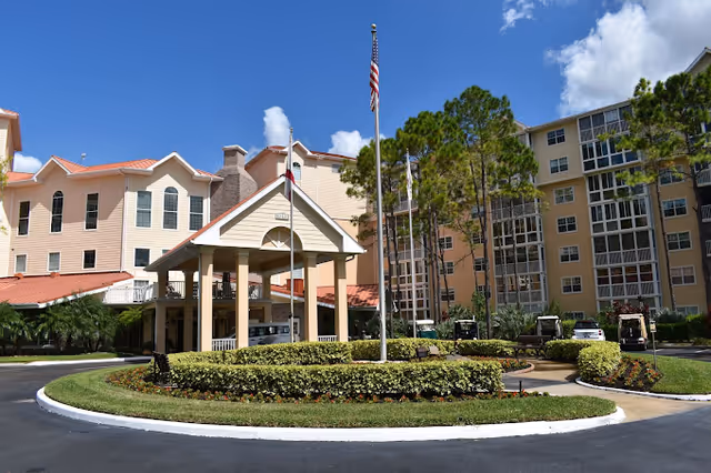 Exterior view of a senior living facility named Freedom Plaza with a circular driveway, landscaped greenery, and an American flag on a flagpole in front of the building under a blue sky with some clouds.