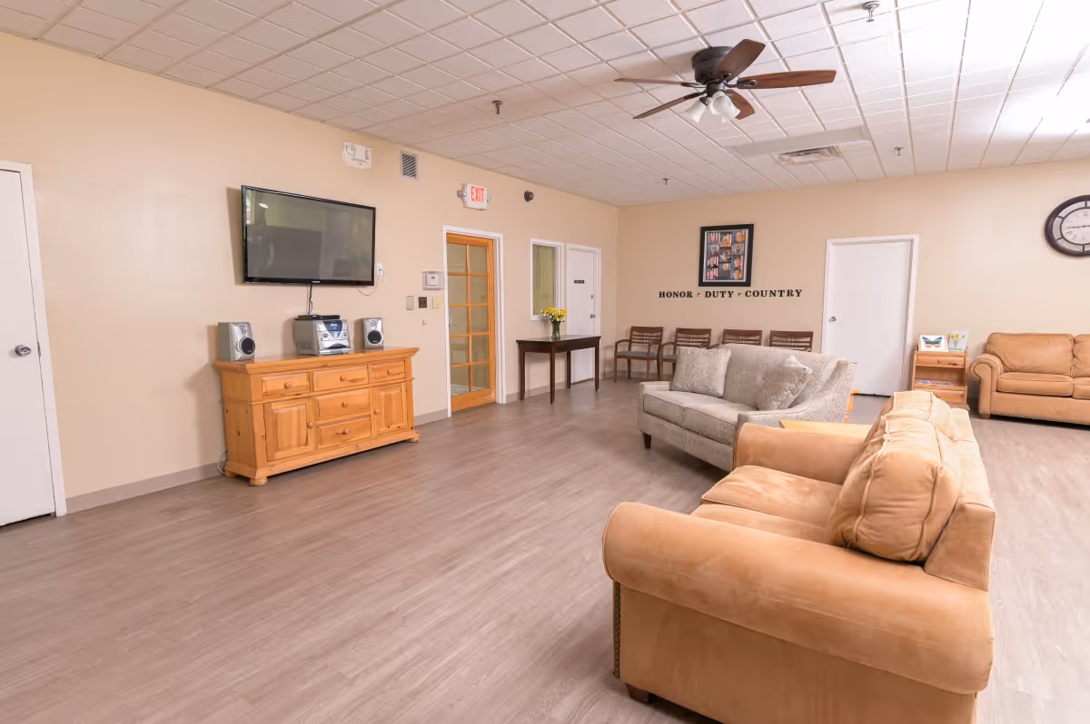 A spacious living room area in a senior living facility with beige walls and wood flooring. The room features multiple sofas and chairs arranged for seating, a wooden cabinet with a stereo system and a flat-screen TV mounted above it. There is a ceiling fan with lights, a clock on the wall, and a framed collage with the words 'HONOR DUTY COUNTRY' beneath it. Several doors and a small table with flowers are also visible.