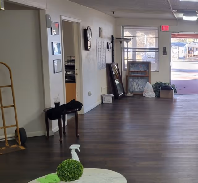 Interior common area with dark wood floors, a small round table and plant in the foreground, leaning framed pictures and a clock along the wall, and an exit door to the outside.