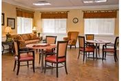 A cozy common area with several round tables and chairs arranged on a wood-patterned floor. There is a patterned sofa and an armchair near the windows, which have brown valances. The walls are light-colored, and a clock is mounted above the windows.