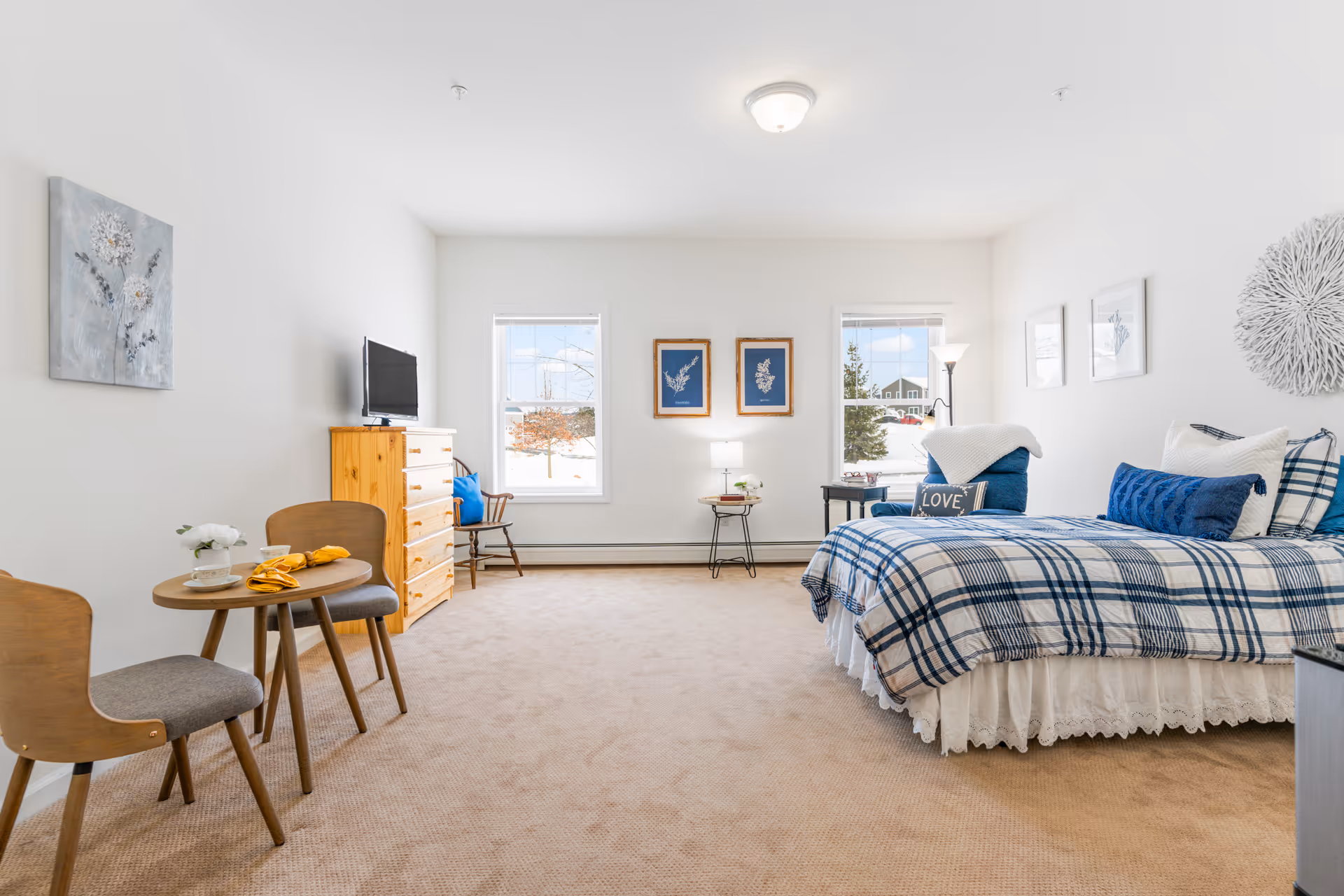A bright and spacious bedroom with beige carpet, a bed with blue and white plaid bedding, two windows letting in natural light, a small round table with two chairs, a wooden dresser with a TV on top, and various wall decorations including framed pictures and a textured white circular piece.