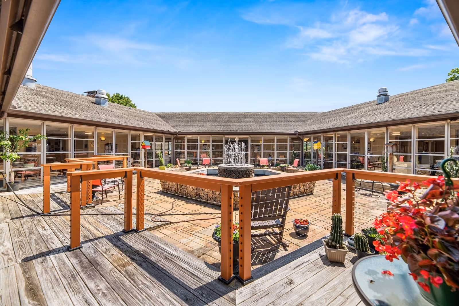 Outdoor courtyard area with a central stone fountain surrounded by a tiled patio and wooden deck. The courtyard is enclosed by a building with large windows and a shingled roof. There are several chairs and potted plants, including red flowers and cacti, placed around the space under a clear blue sky.
