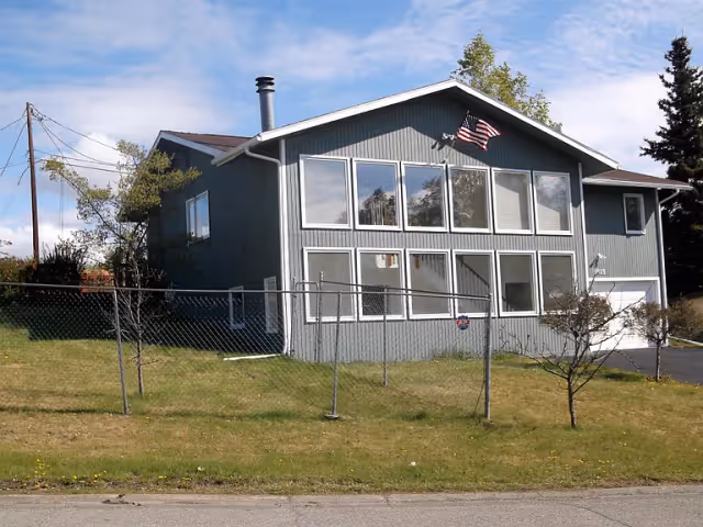 Exterior view of a two-story gray building with multiple windows and an American flag mounted near the roof peak. The building is surrounded by a grassy lawn with a chain-link fence in front and a few small trees. The sky is partly cloudy.
