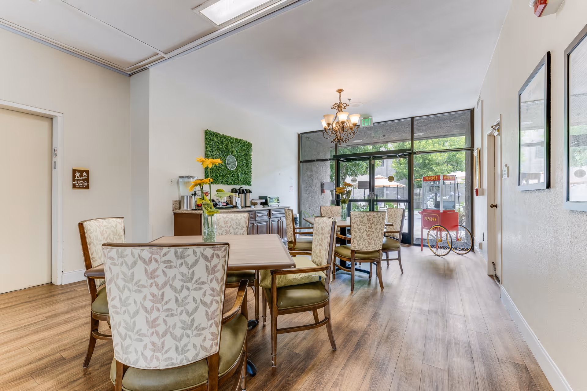 Interior view of a dining area in a senior living facility with wooden floors, several tables and chairs with floral upholstery, a chandelier, a refreshment station with coffee and water dispensers, and a popcorn machine near glass doors leading outside.