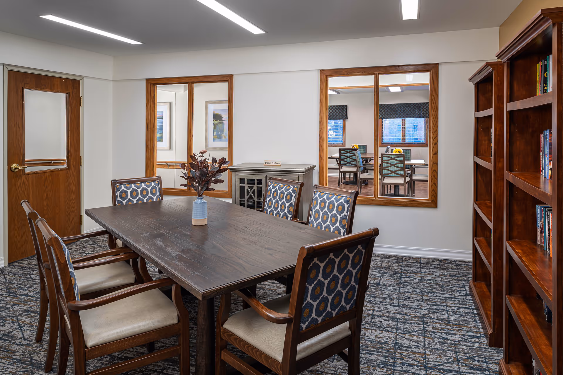 A cozy meeting or reading room with a dark wooden table surrounded by six cushioned chairs with patterned backs. A small vase with decorative branches is placed on the table. The room has a carpeted floor with a geometric pattern, white walls, and wooden framed windows looking into another room with tables and chairs. There is a wooden bookshelf on the right side filled with books and a wooden door with a frosted glass panel on the left.