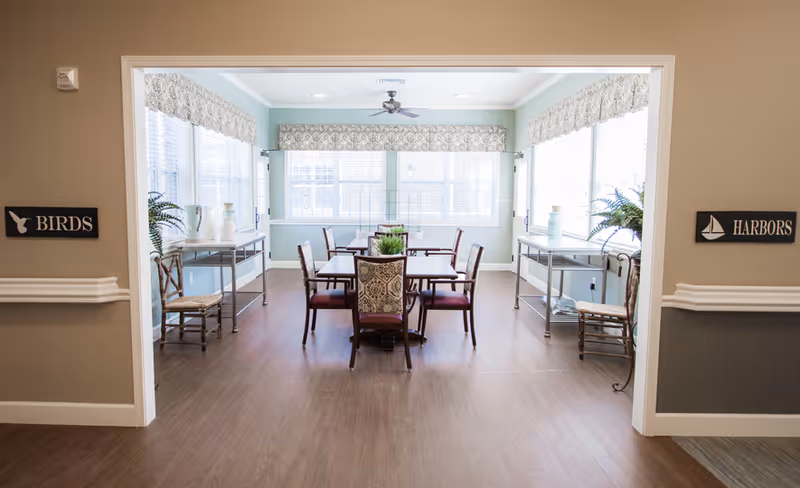 A bright dining room with a rectangular table surrounded by eight chairs, two metal carts with decorative vases and plants on either side, large windows with patterned valances letting in natural light, and signs on the walls labeled 'BIRDS' and 'HARBORS'.