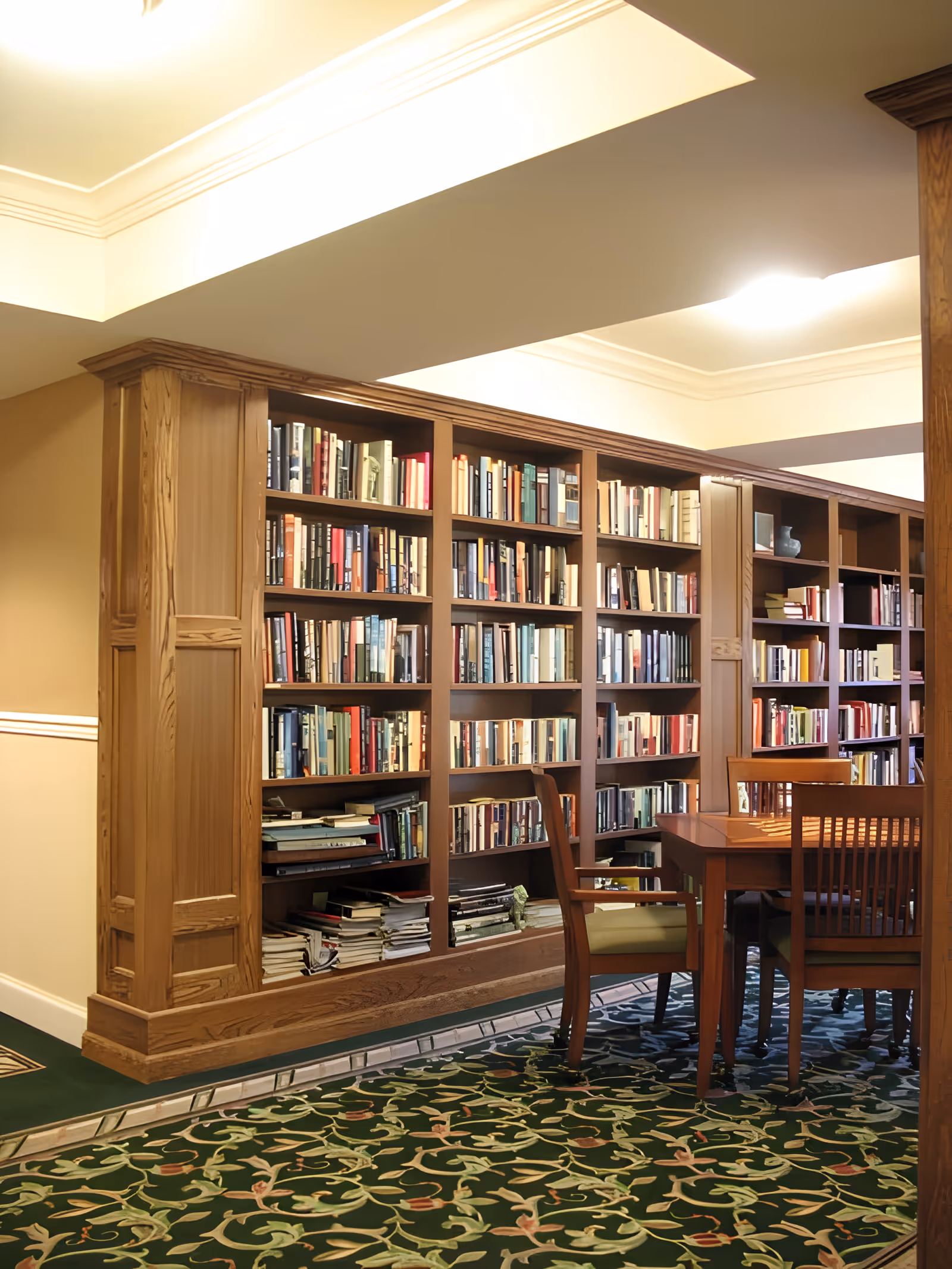 A cozy library room with wooden bookshelves filled with books and a wooden table surrounded by chairs on a green patterned carpet.