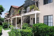 Exterior view of a two-story senior living apartment building with balconies, stairs, and lush green landscaping including bushes and trees under a clear sky.