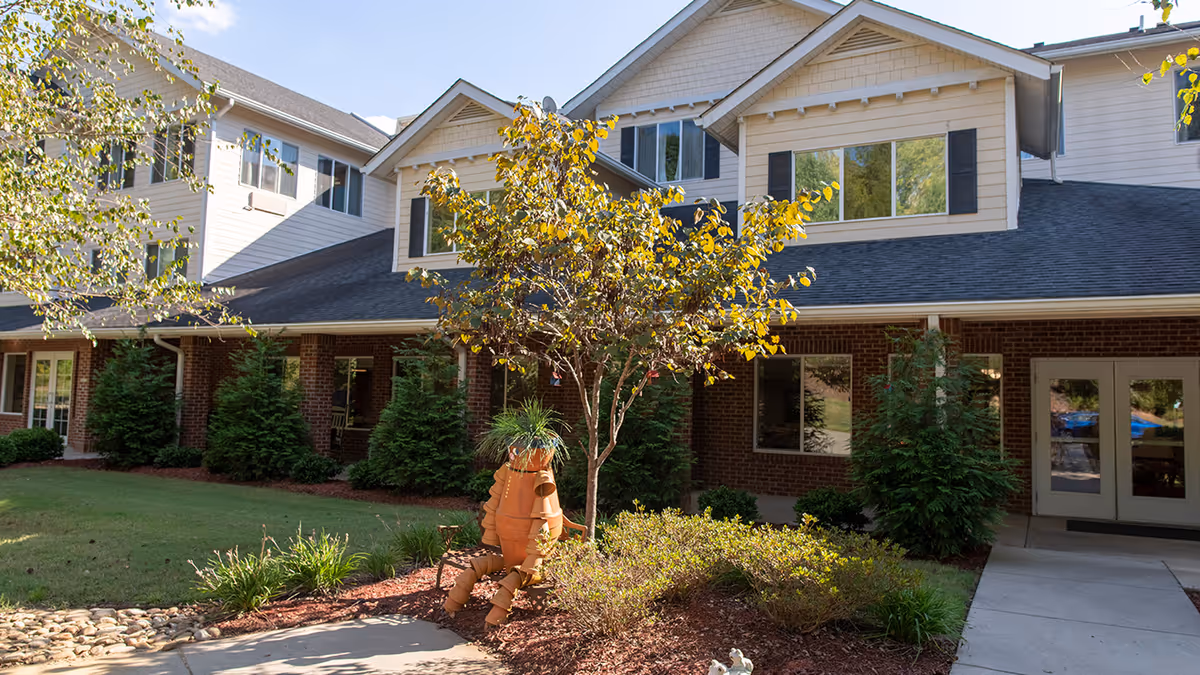 Exterior view of a senior living facility building with brick and light-colored siding. There is a small landscaped area with bushes, a tree, and a decorative figure made of terracotta pots shaped like a person sitting on a bench. The sky is clear and blue.