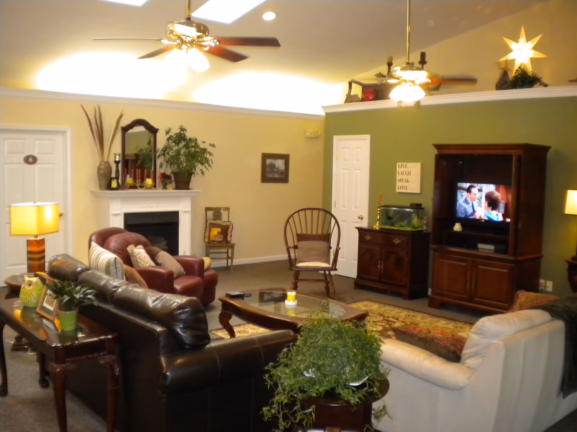 A cozy living room in an assisted living facility with a black leather sofa, a white armchair, a red armchair, a wooden rocking chair, a coffee table, and a TV cabinet with a television showing a scene from a movie. The room has a fireplace with decorative items on the mantel, plants, lamps, ceiling fans, and a green accent wall with a motivational sign.