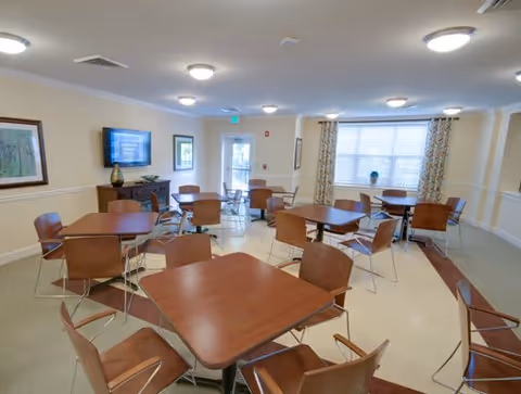 A well-lit dining room with multiple square wooden tables and matching chairs arranged neatly. The room has beige walls with white trim, a flat-screen TV mounted on one wall, framed artwork, large windows with patterned curtains, and a door leading outside.