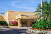 Entrance of a beige single-story building with a covered porte-cochère, palm trees and landscaped flower beds.