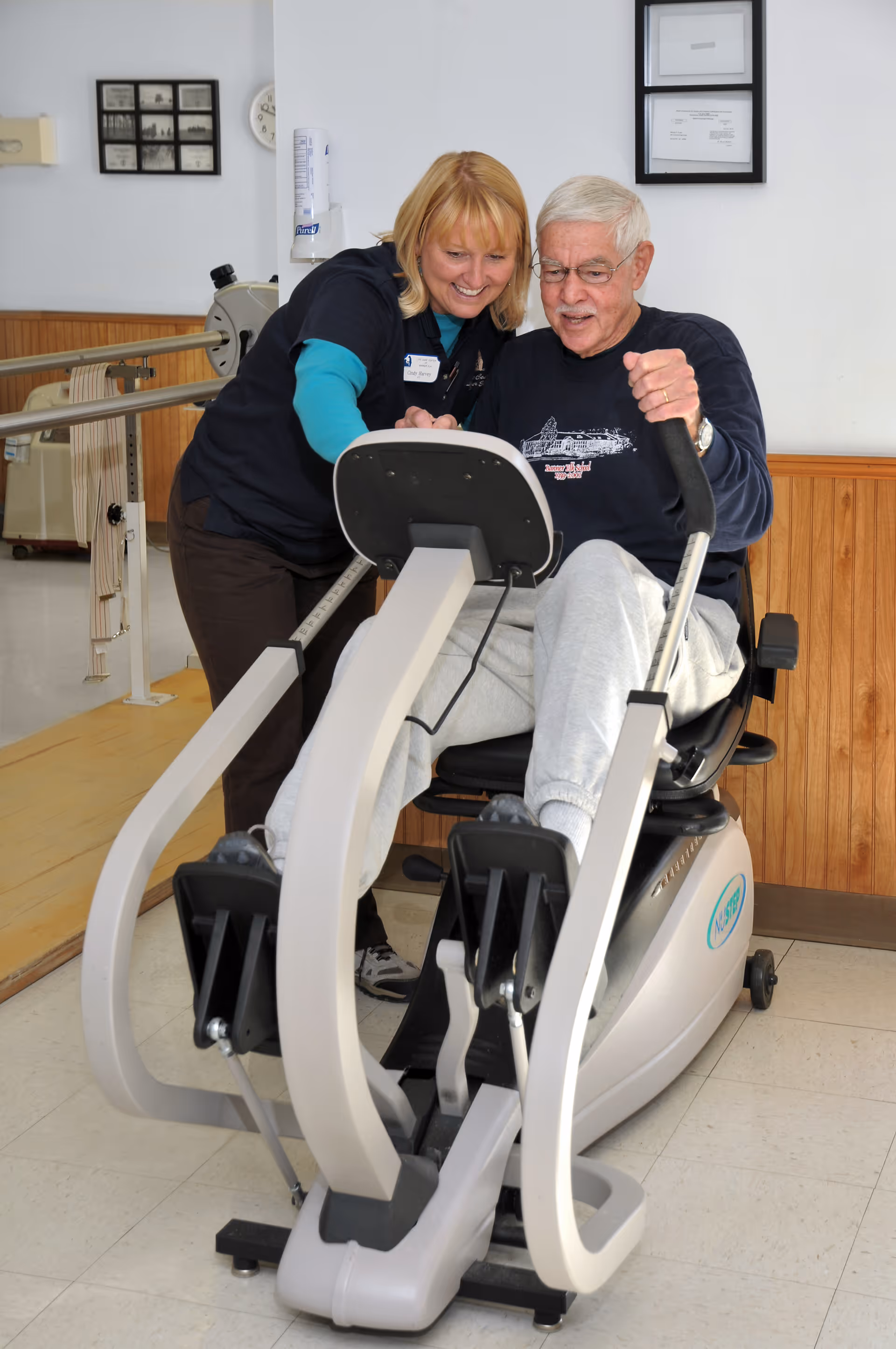 An elderly man using a seated exercise machine with the assistance of a female staff member in a rehabilitation or fitness room. The woman is smiling and pointing at the machine's display while the man pedals with his feet. The room has light-colored walls and wood paneling along the lower half.