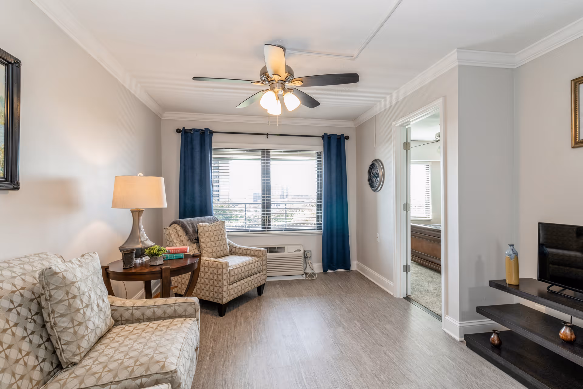 A cozy living room with two patterned armchairs, a wooden side table with a lamp and books, a ceiling fan with lights, a large window with blue curtains, and a TV on a dark wooden stand. The room has light-colored walls and wood flooring, with an open doorway leading to a bedroom.