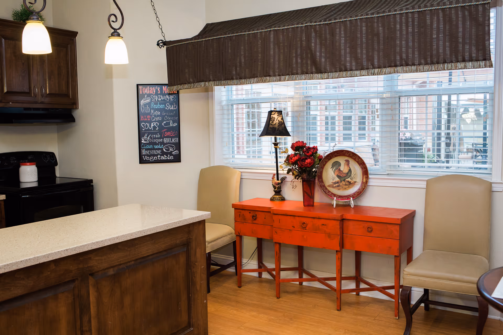 Interior view of a kitchen area with a wooden island countertop, a black stove, and dark wooden cabinets. Two beige chairs flank a red console table adorned with a lamp, a vase of red flowers, and a decorative plate with a rooster design. A window with white blinds and a brown valance is behind the console table. A chalkboard menu is mounted on the wall listing sandwiches and soups.
