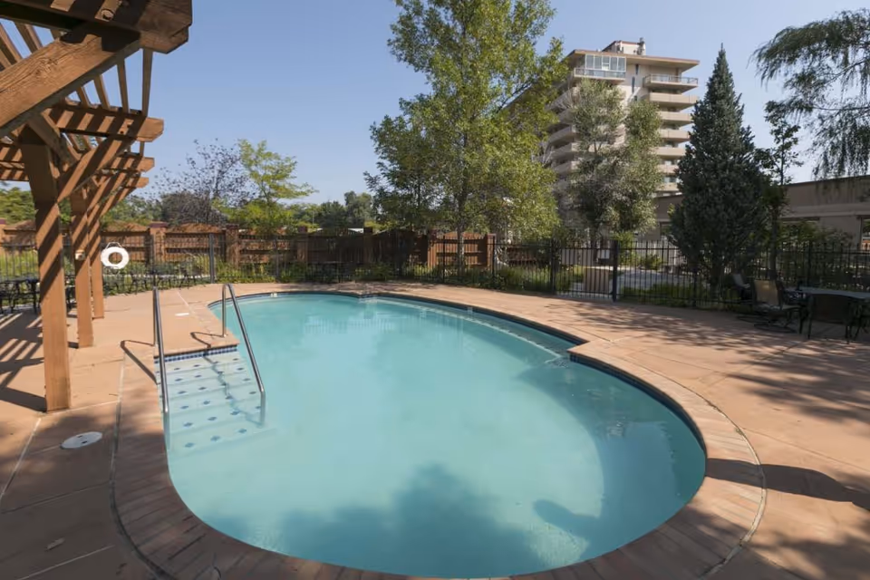 Outdoor swimming pool with clear blue water surrounded by a concrete deck, metal handrails, and a wooden pergola on the left side. Trees, shrubs, and a multi-story building are visible in the background under a clear blue sky.