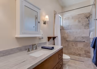 Bathroom with a light-colored stone countertop and wooden vanity with a sink, a wall-mounted mirror cabinet, a wall sconce light, a towel rack with a dark blue towel, and a walk-in shower with beige stone tiles and a grab bar.