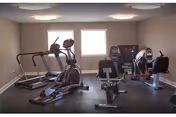 A small fitness room with exercise equipment including two treadmills, an elliptical machine, a stationary bike, and a recumbent bike. The room has beige walls, two windows letting in natural light, and ceiling lights.