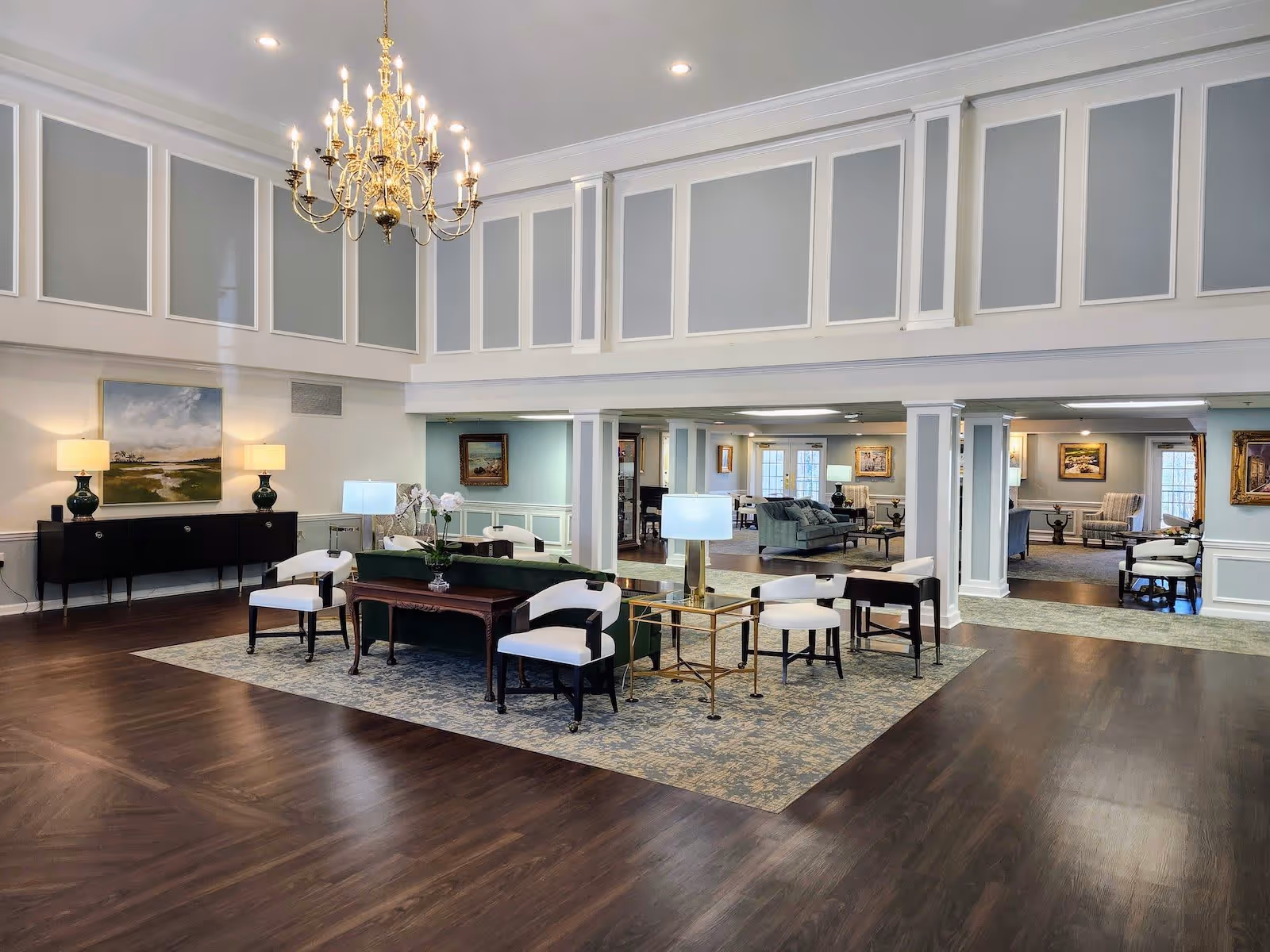 Spacious and elegant senior living facility common area with high ceilings, a large gold chandelier, and soft lighting. The room features a seating arrangement with white cushioned chairs around a dark wood table on a patterned rug. Additional seating areas with sofas and armchairs are visible in the background, along with framed artwork on the walls and decorative lamps on side tables.