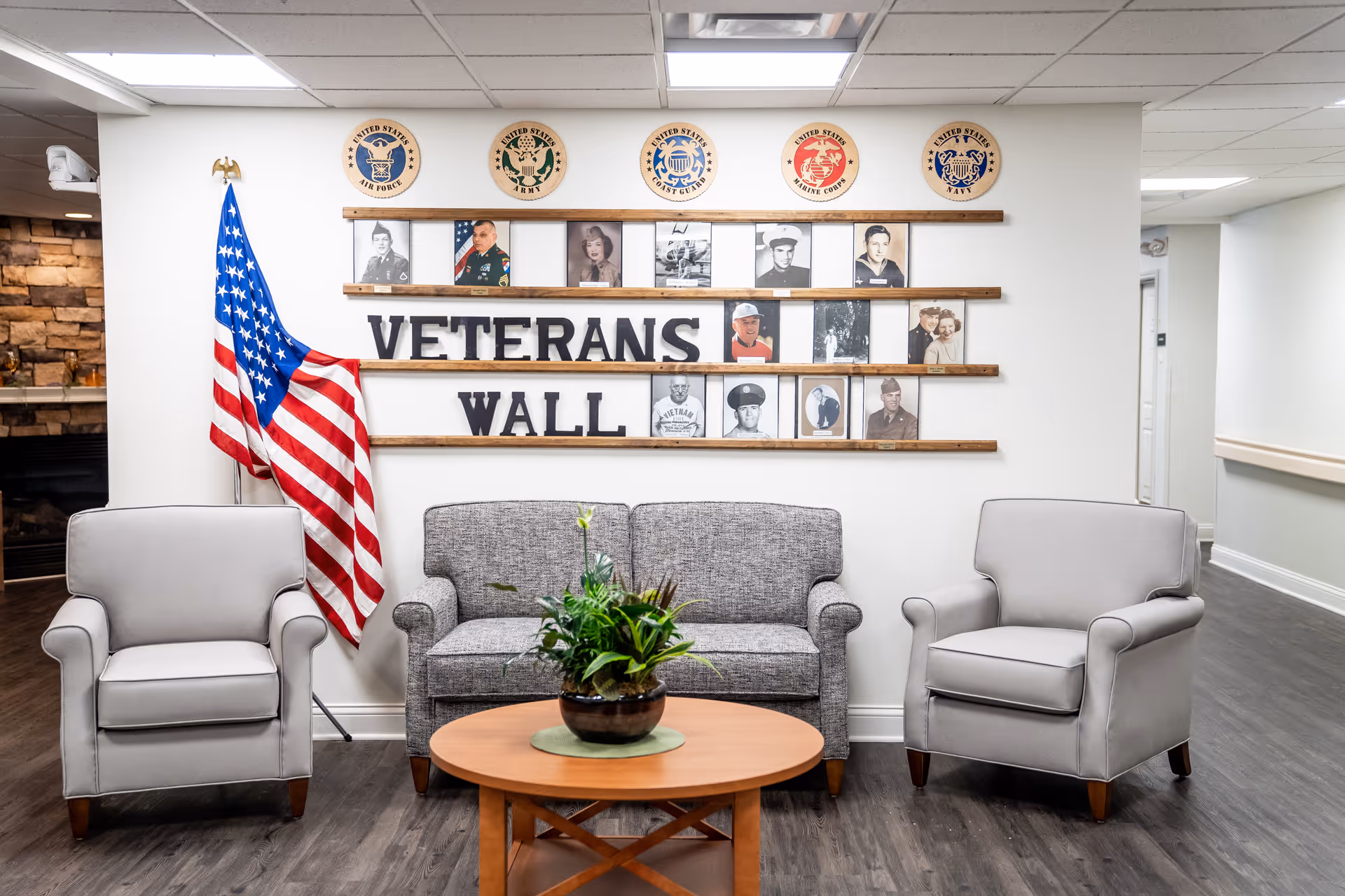 Seating area with a "Veterans Wall" of photos and military emblems, an American flag, a sofa and two armchairs around a coffee table with a plant.