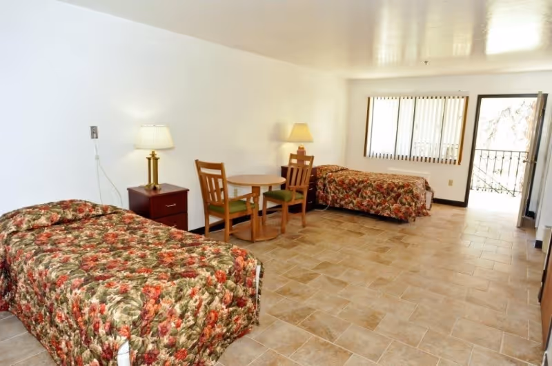 A spacious room with two single beds covered in floral bedspreads, a small round wooden table with two chairs, two bedside tables each with a lamp, a window with vertical blinds, and an open door leading outside.