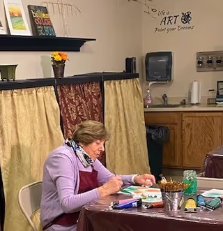 An elderly woman wearing a purple sweater and a red apron is seated at a table covered with a brown plastic tablecloth, painting on a small canvas. The room has beige walls with a paper towel dispenser and some cabinets in the background. There is a decorative curtain hanging under a shelf with books and a small vase with flowers. On the wall, there is a decal that reads 'Life is ART Paint your Dreams' with a butterfly graphic.
