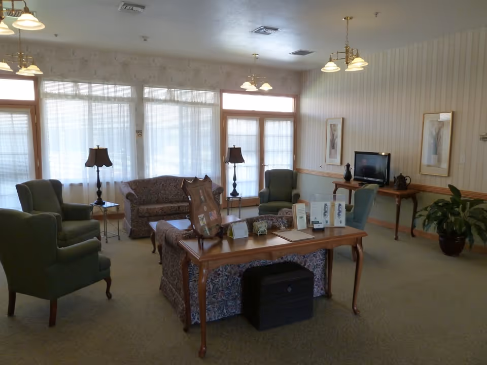 A cozy living room area with patterned sofas, green armchairs, a wooden coffee table with informational brochures, two table lamps, a TV on a wooden console, framed artwork on the walls, and large windows with sheer curtains letting in natural light.