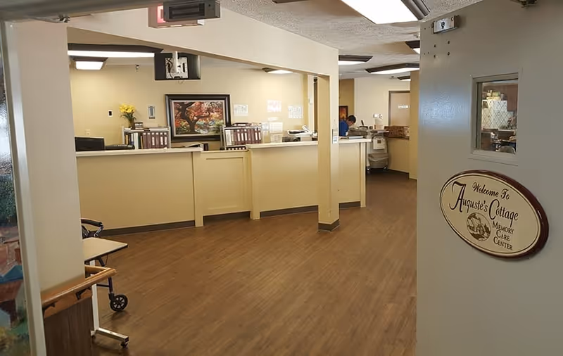 Interior view of a reception or nursing station area in a senior living facility with a wooden floor, beige walls, a counter with office supplies, a painting on the wall, and a sign on the door that reads 'Welcome To Auguste's Cottage Memory Care Center'.