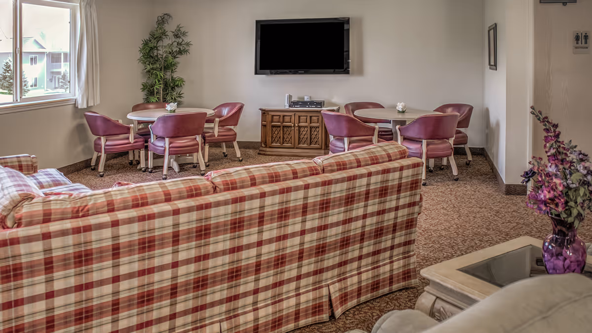 A cozy senior living common room with a plaid sofa in the foreground, round tables with pink chairs, a wall-mounted TV, and decorative plants.