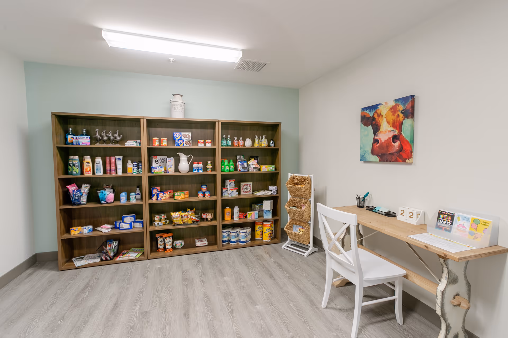 A well-lit room with a wooden shelving unit filled with various snacks, toiletries, and household items. To the right, there is a wooden desk with a white chair, some stationery, and decorative items on top. A colorful painting of a cow hangs on the wall above the desk. The floor has light gray wood-style flooring.