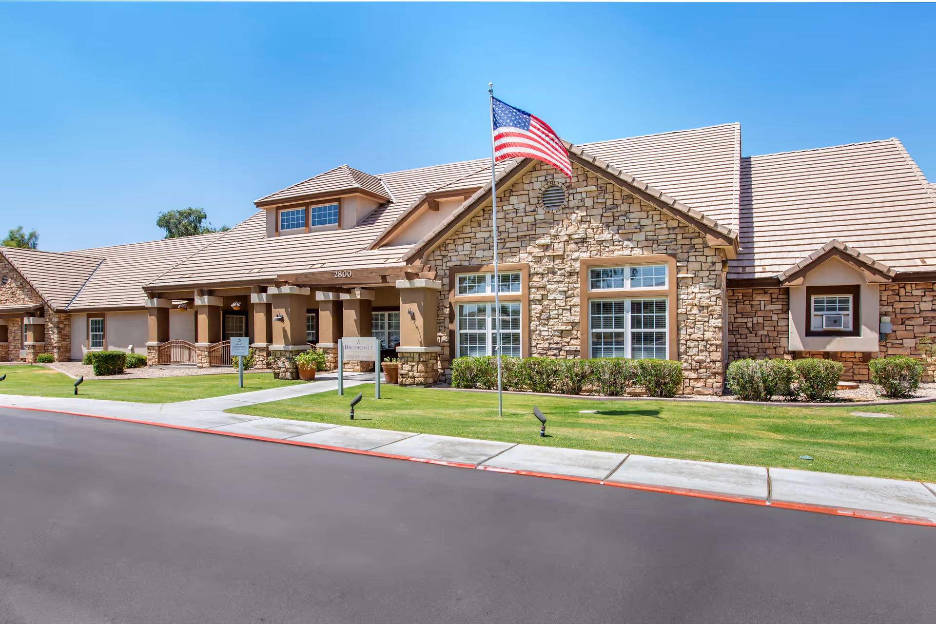 Exterior view of Brookdale Chandler Ray Road facility showing a single-story building with stone and stucco facade, a tiled roof, large windows, an American flag on a flagpole, and a well-maintained lawn with shrubs and a sidewalk.