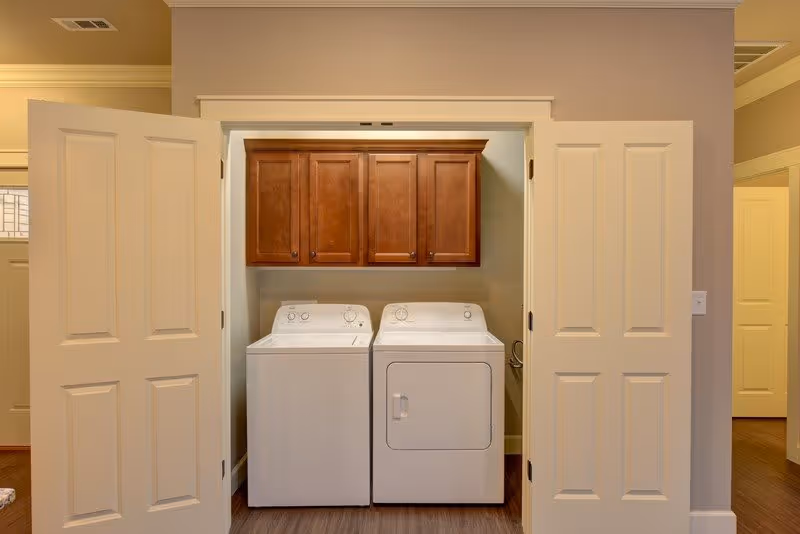 A laundry area with a white washing machine and dryer side by side, enclosed by double white doors. Above the machines, there are wooden cabinets for storage. The walls are painted beige and the floor has a wood finish.