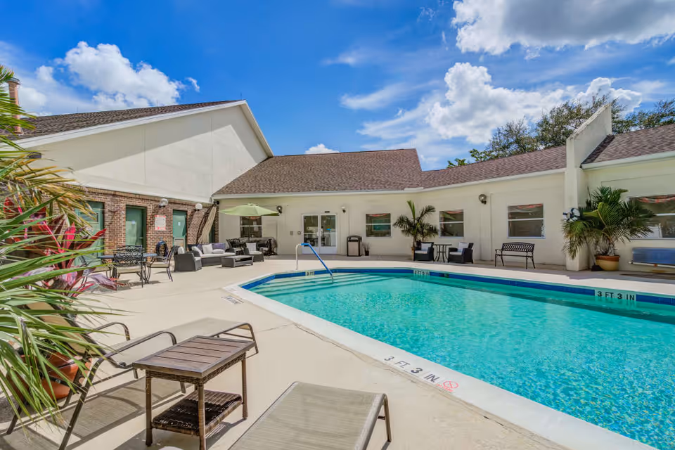 Outdoor swimming pool area at Merrill Gardens at Barkley Place with lounge chairs, tables, umbrellas, and potted plants surrounding the pool under a partly cloudy blue sky.