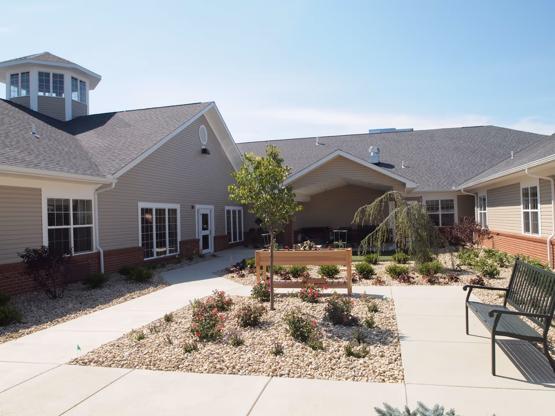 Outdoor courtyard area of The Willows at Willard senior living facility featuring a landscaped garden with small trees, bushes, and flowers surrounded by a paved walkway. The building has beige siding with white trim and a covered seating area in the background. A black metal bench is visible on the right side of the walkway.