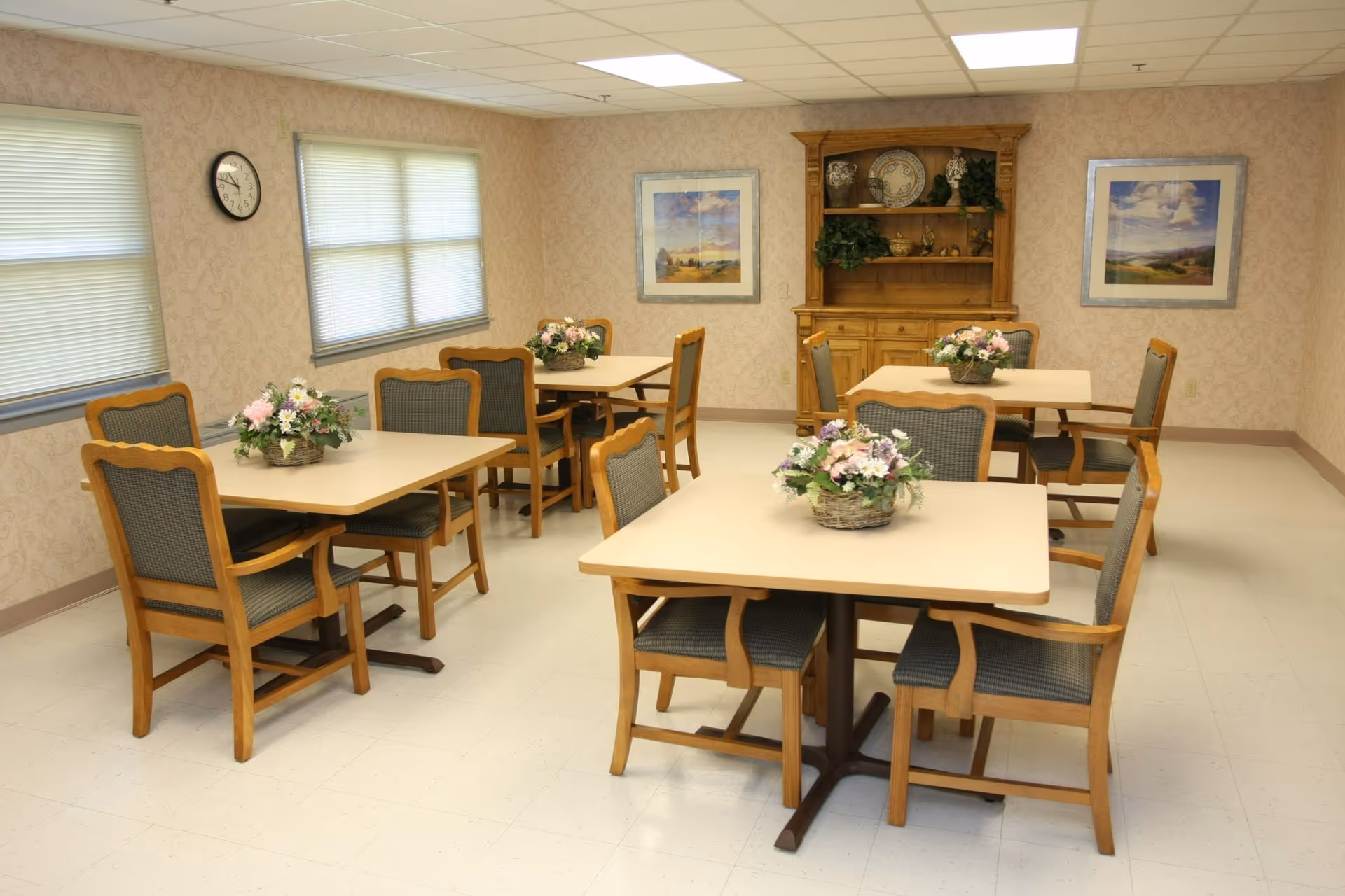 A dining room with four square tables, each surrounded by four wooden chairs with cushioned seats and backs. Each table has a floral centerpiece in a basket. The room has beige patterned wallpaper, two windows with blinds, a clock on the wall, two framed landscape paintings, and a wooden hutch with decorative items and greenery.