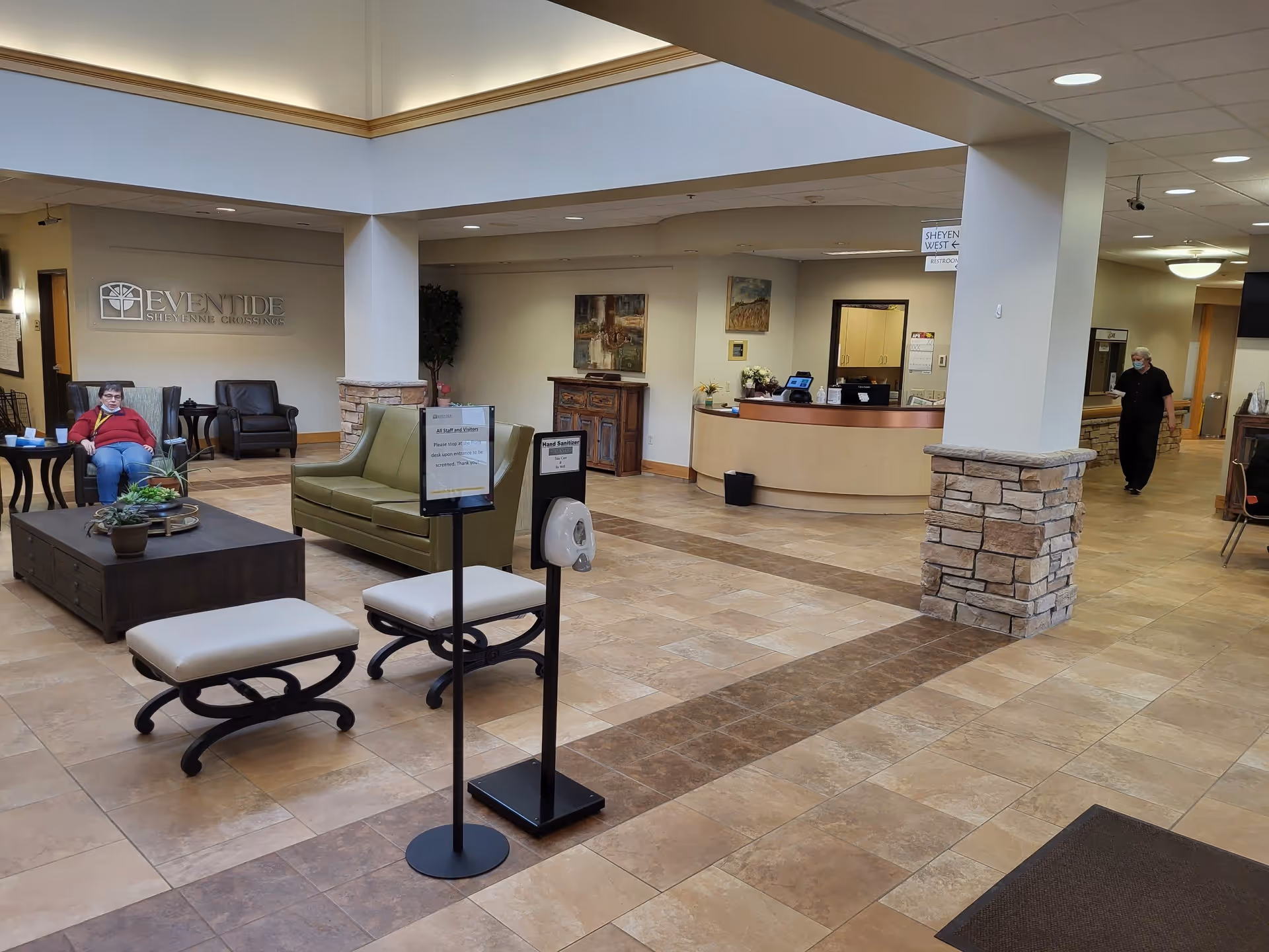 A spacious senior living facility lobby with seating, a reception desk, and a wall sign reading 'Eventide Sheyenne Crossings'.