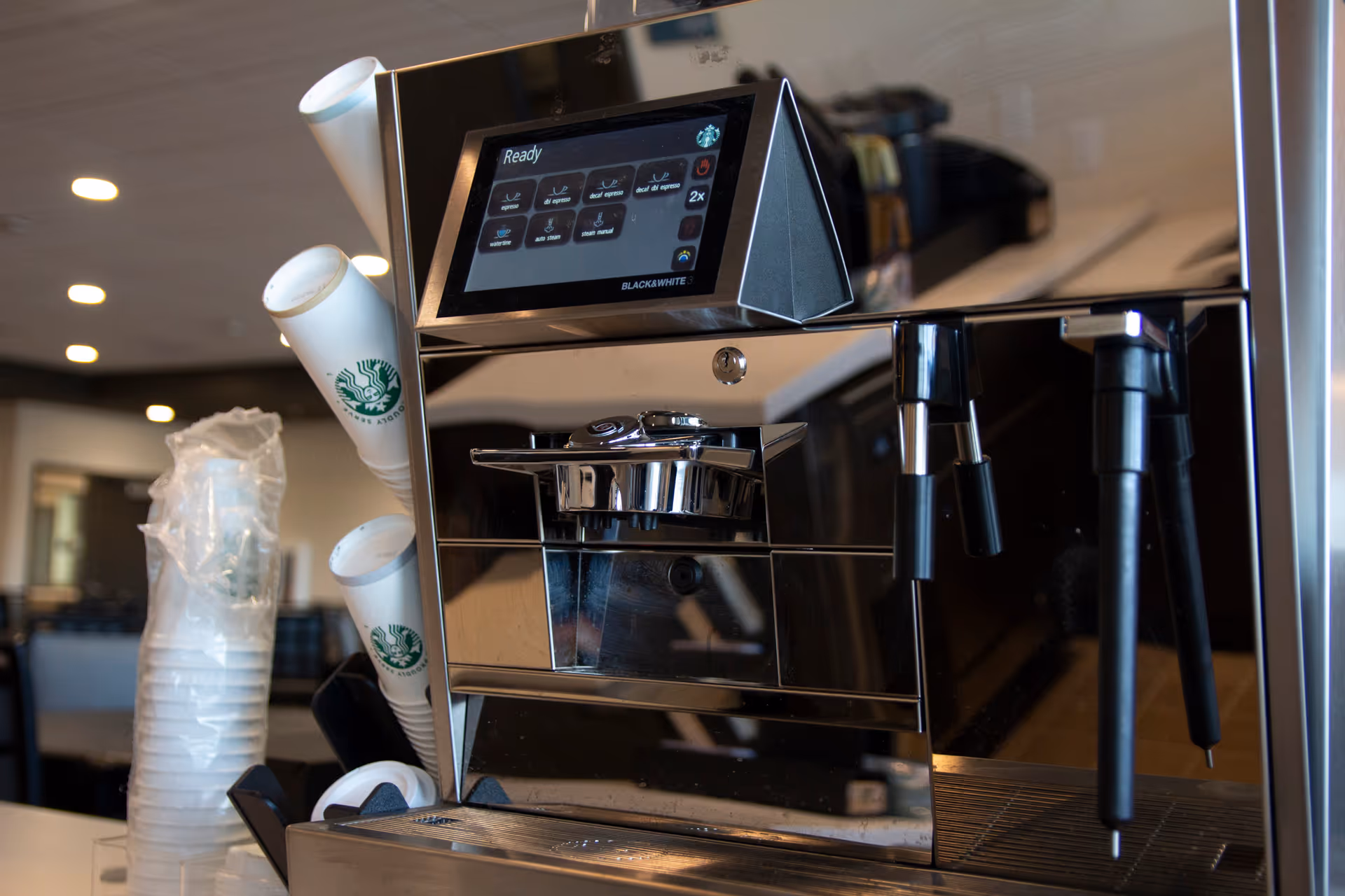 Commercial espresso machine with stacked Starbucks cups on a countertop in a dining area.