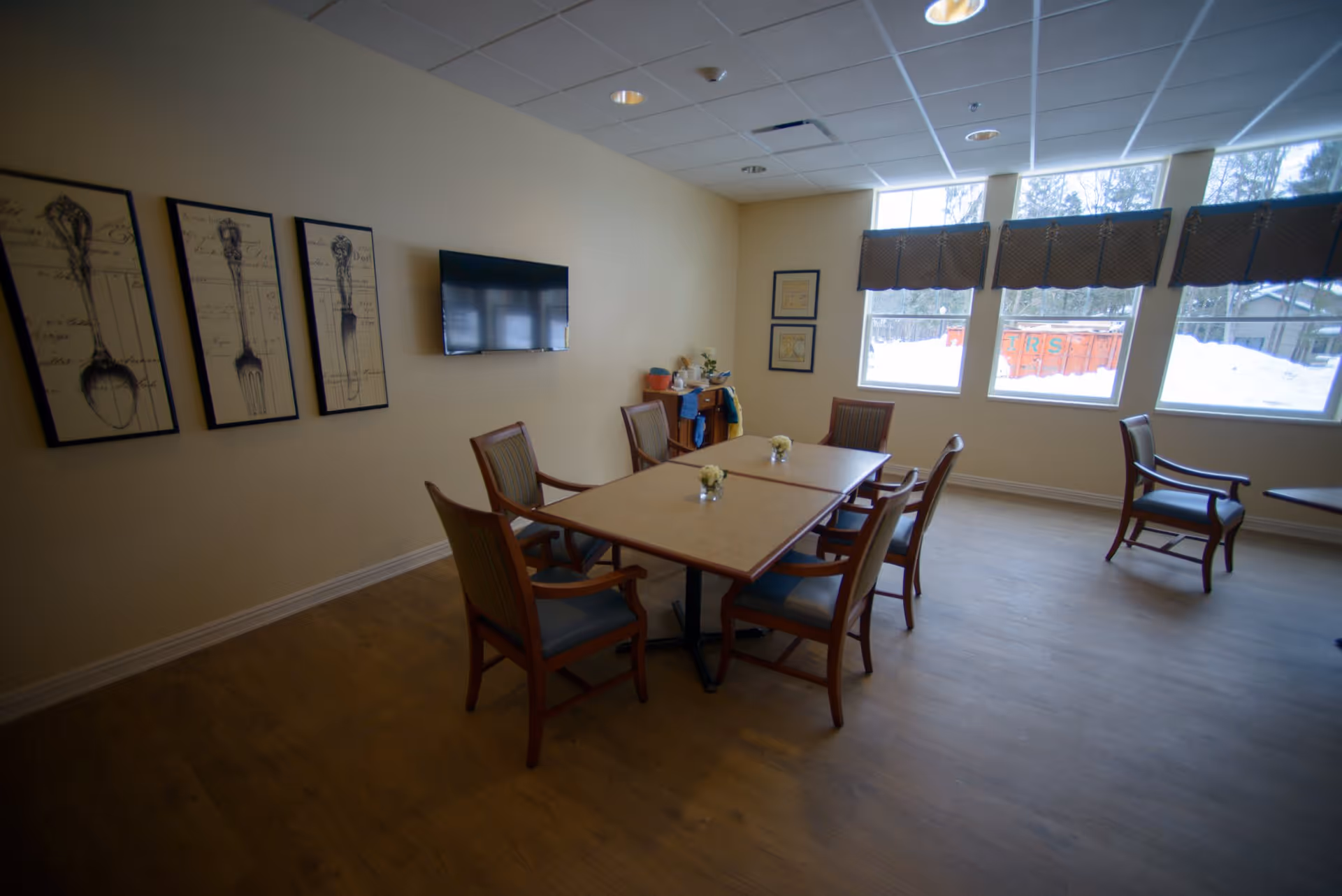 A dining room with a rectangular table surrounded by six wooden chairs with cushions. The room has three large windows with valances, letting in natural light and showing a snowy outdoor scene. On the wall, there are three framed pictures of vintage silverware and a flat-screen TV mounted. The floor is wooden, and there are small flower arrangements on the table.